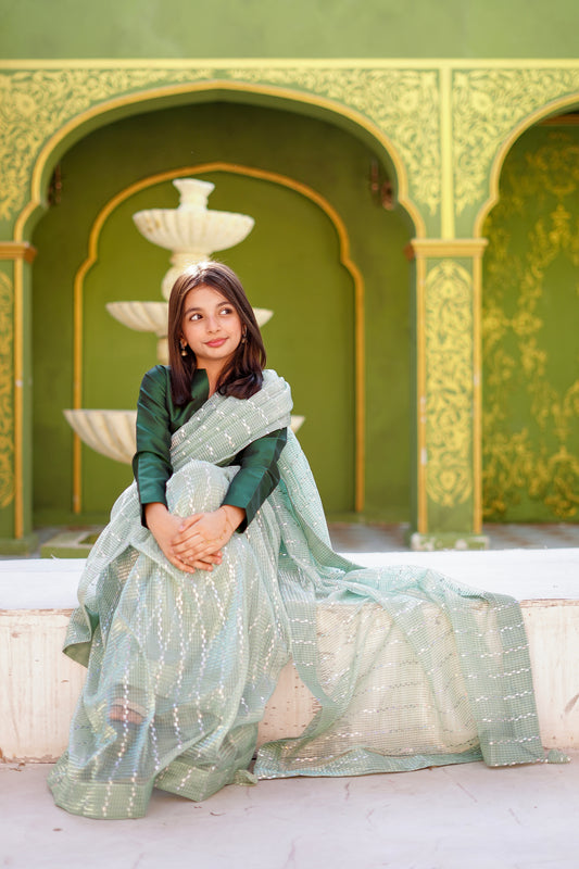 Kid in a light green saree sitting in an ornate outdoor setting with green walls and arches.