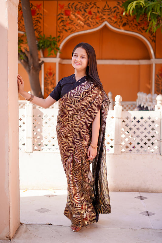 Kid in a black and golden saree standing in front of an ornate archway.