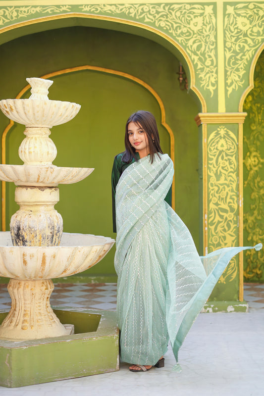 Kid in a light green saree standing next to a marble fountain with decorative arches in the background.